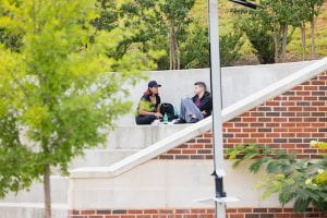 Two students sitting at a distance on steps, lounging in comfortable conversation, surrounded by trees.
