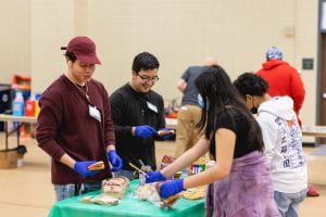 Group of casually dressed students standing around a table wearing blue gloves are smiling and spreading peanut butter on bread to make sandwiches.