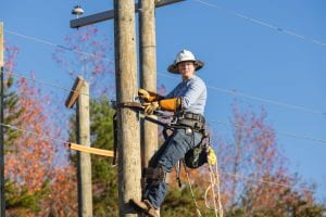 Duke Energy employee in a hard hat is climbing a telephone pole while wearing a safety harness. She is wearing heavy gloves and special boots.