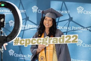 Student Cristina Vargas stands in her gray cap and gown against a background with the Central Piedmont logo, holding a sign that reads #cpccgrad22