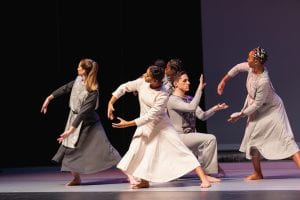A group of dancers on stage in plain frocks, stand with their legs apart and interacting with each other with their arms held in circles.