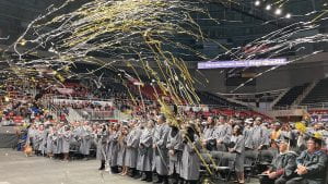 silver and gold streamers fly through the air above graduates heads in the arena.
