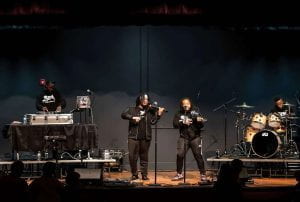 Two black violinists with a DJ and another band member at a drum set on stage.
