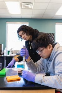 Two students look over work in a lab, holding a pipette and a test tube.