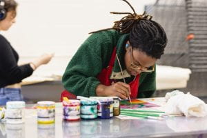 Student painting on a canvas with jars of paint on the table.